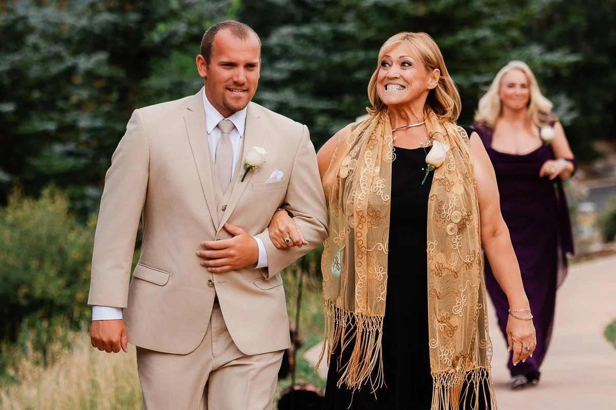 A smiling woman in a black dress and golden scarf walks arm-in-arm with a man in a beige suit. Another woman follows. The scene is joyful and outdoor.