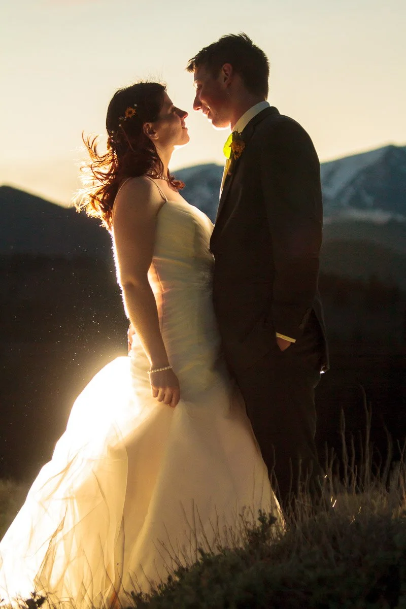 A bride and groom face each other, smiling warmly, backlit by a setting sun. Mountains in the background add a romantic and serene atmosphere.
