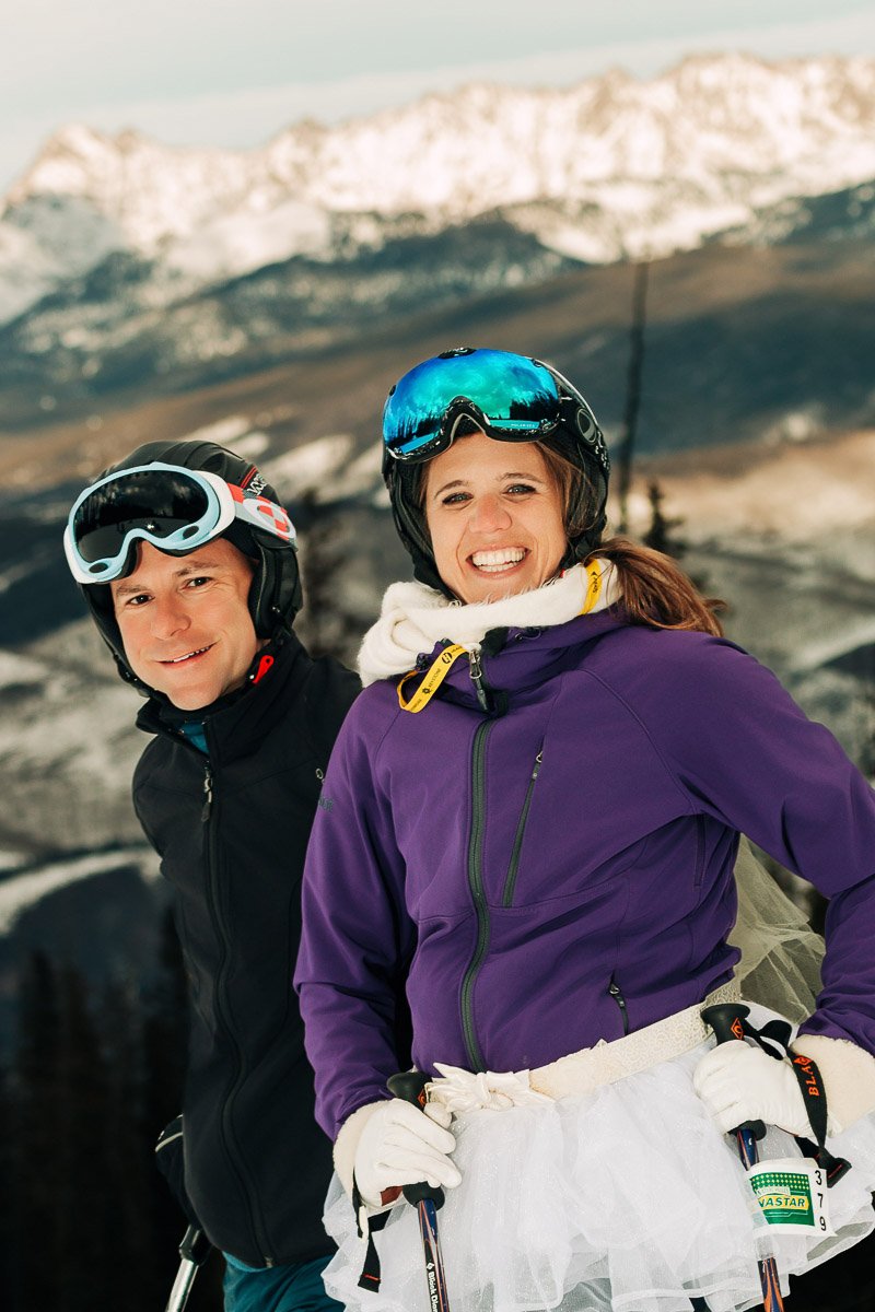 Smiling man and woman in ski gear stand on a snowy mountain slope. The woman wears a purple jacket and white tutu. Snowy peaks and clear sky in the background.