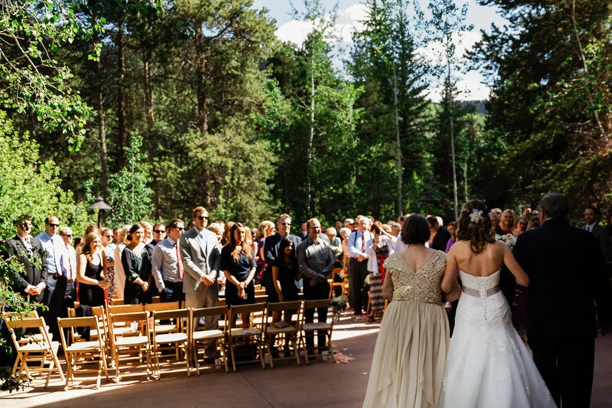 Bride walking down an outdoor forest aisle with parents, surrounded by standing guests in daylight. The scene feels joyful and serene.