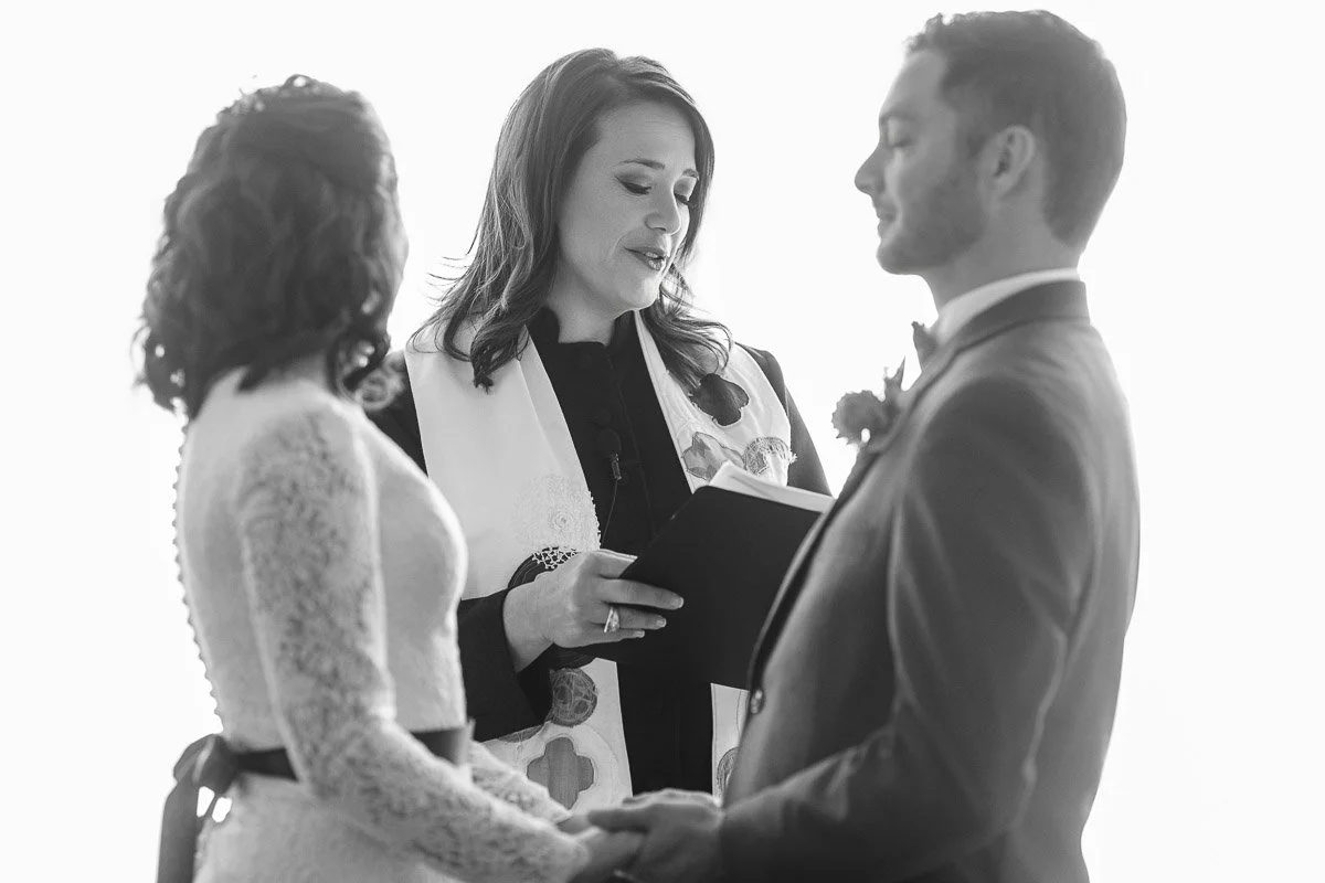 A couple holds hands, facing each other during a wedding ceremony, as the officiant reads from a book. The setting is formal and intimate.