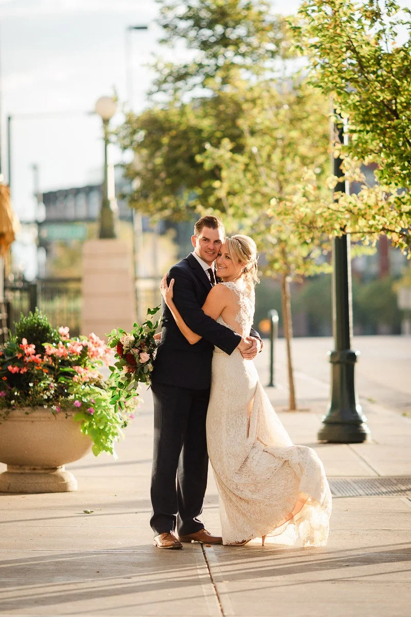 A couple in wedding attire embraces on a sunlit sidewalk. She holds a bouquet, and they smile warmly, surrounded by trees and flower planters. Romantic and joyful.