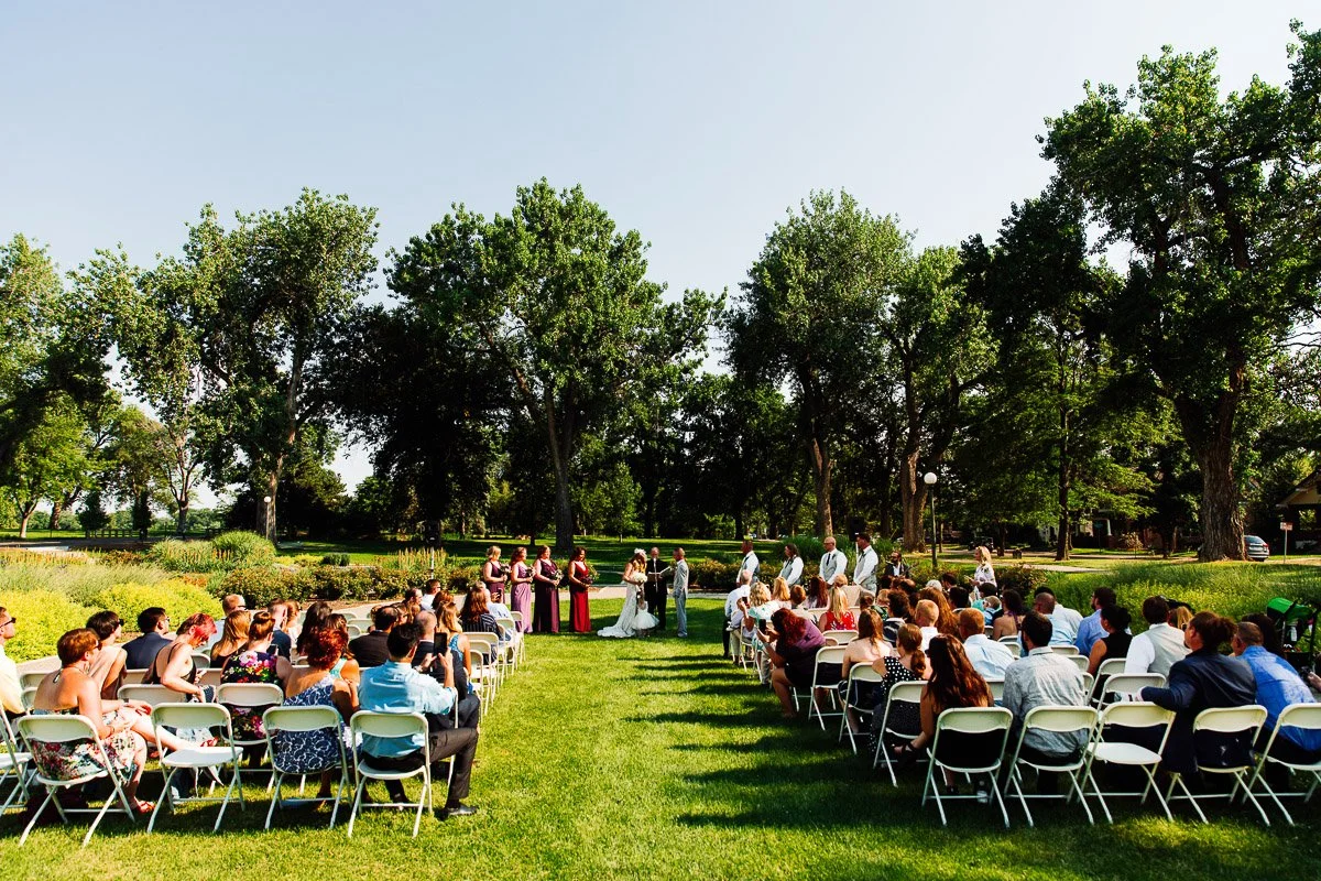 Outdoor wedding ceremony with guests seated on folding chairs in rows, facing a couple and wedding party. Surrounded by lush trees under a clear sky.