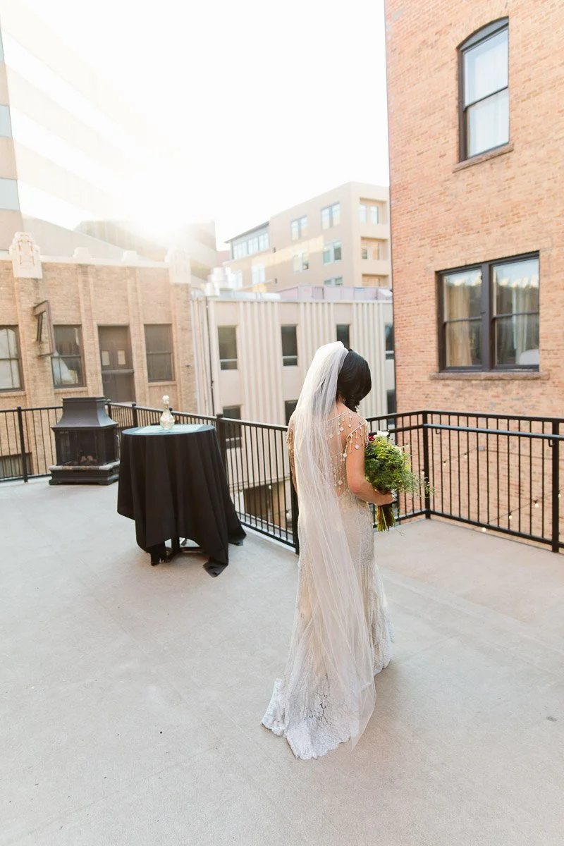 A bride in a flowing white gown and veil stands on a sunlit rooftop terrace, holding a bouquet. The setting has urban buildings, metal railings, and a table.