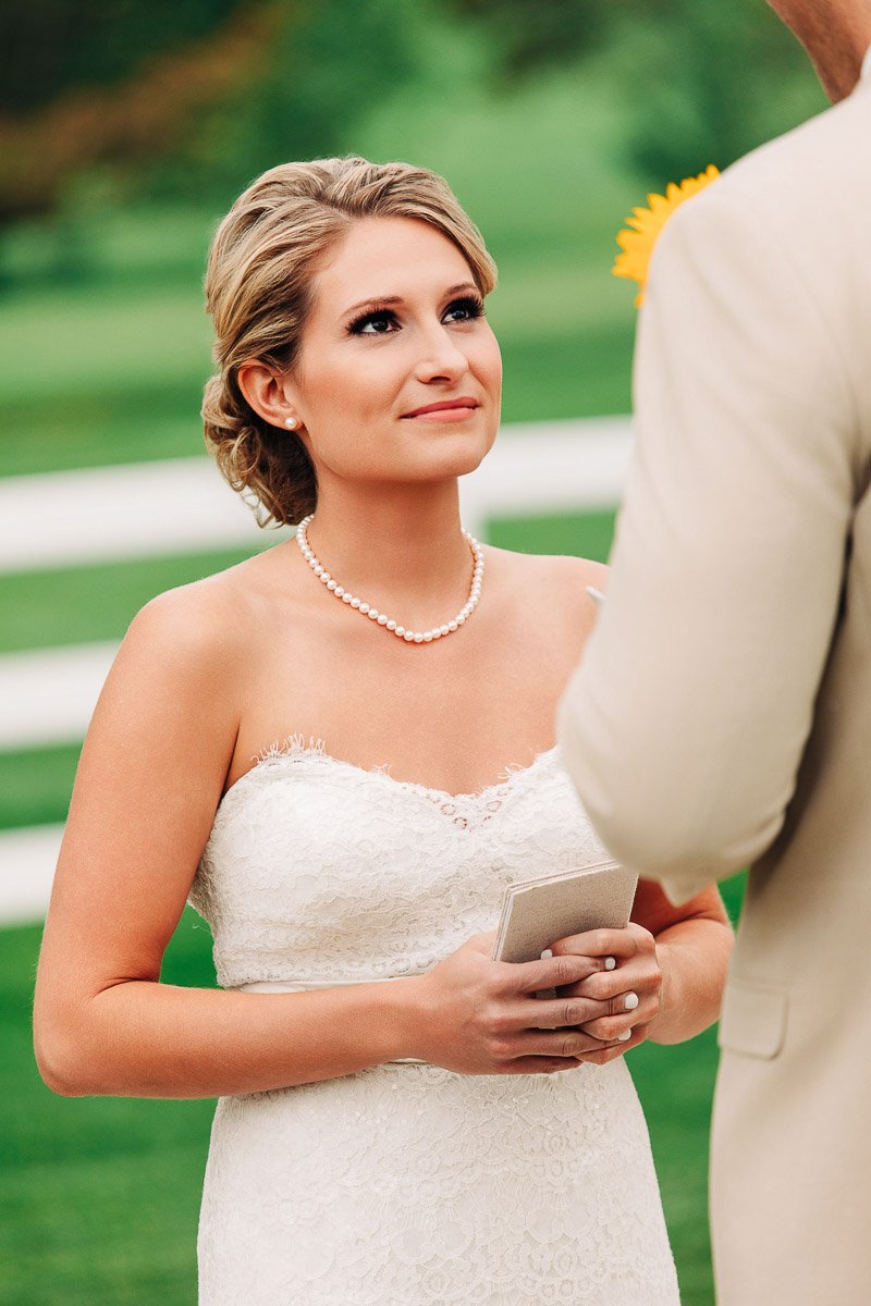 A bride in a lace gown and pearl necklace looks lovingly at another person holding a sunflower. The background features a green lawn and white fence.