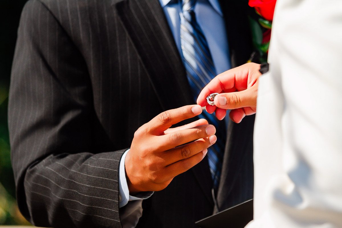 A person in a black pinstripe suit holds out their hand as another hands them a wedding ring. The moment captures a sense of anticipation and intimacy.