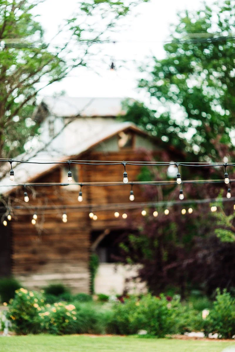 String lights hang in front of a rustic wooden barn surrounded by lush greenery and trees, evoking a serene and tranquil countryside ambiance.