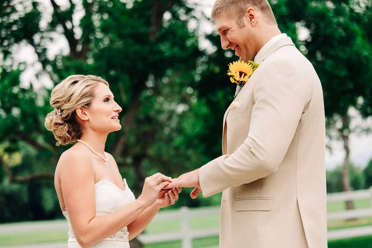 Bride and groom exchange rings in an outdoor setting. The bride, in a white dress, smiles gently, while the groom, in a beige suit with a yellow boutonniere, looks fondly at her. Lush greenery in the background conveys a joyful, romantic mood.