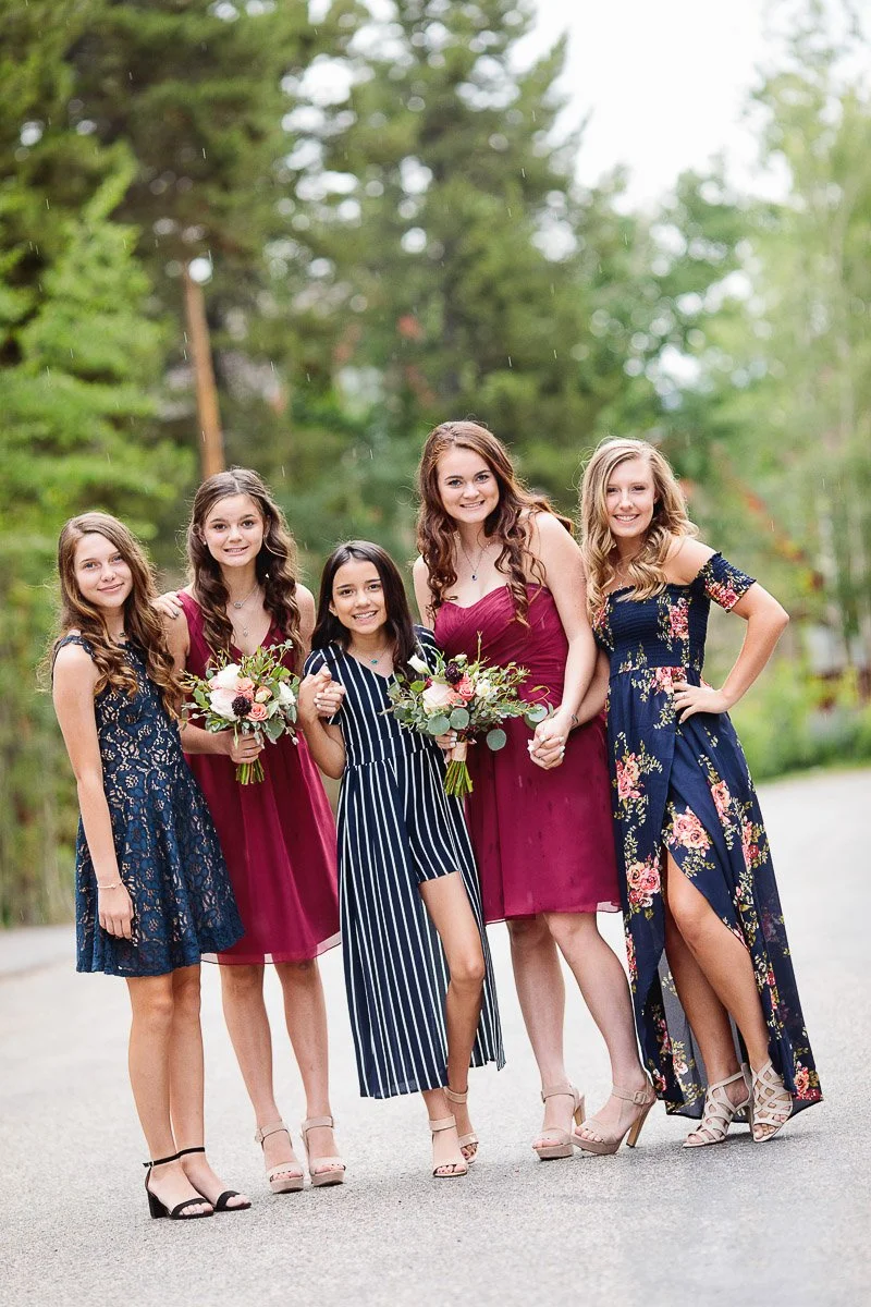 A group of five smiling young girls stands on a road surrounded by greenery. They wear elegant dresses in floral and solid patterns, holding bouquets. The mood is joyful.