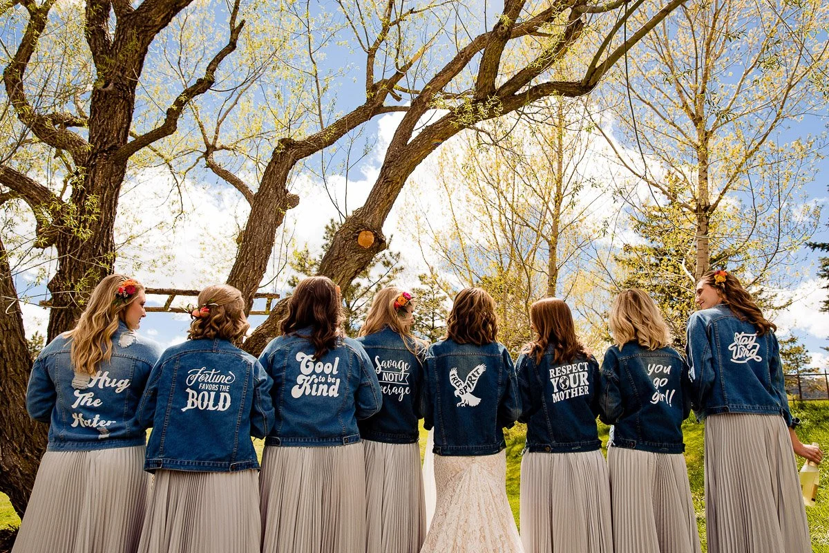 A group of women in denim jackets and gray skirts stand with backs to the camera. Each jacket displays unique empowering messages. Trees and blue sky form the backdrop, exuding a joyful and supportive atmosphere.