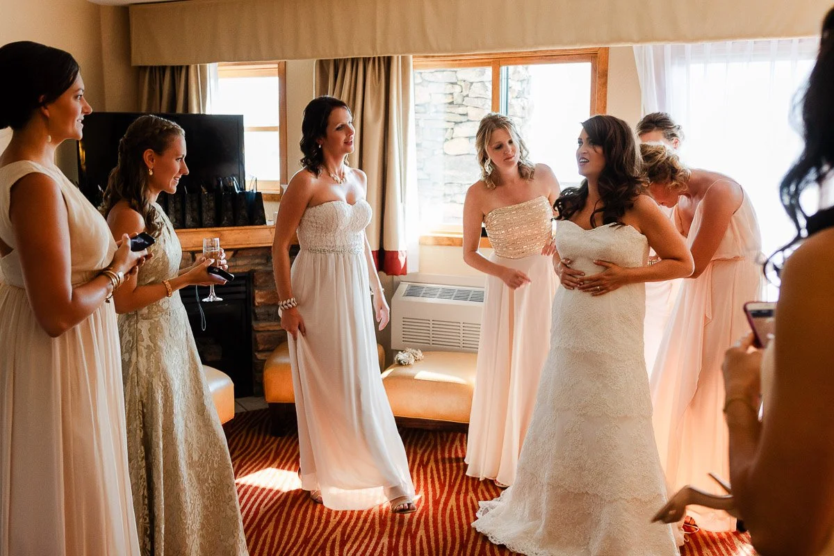 Seven women in elegant dresses share a joyful moment in a warmly lit room. The bride, in white, is the center of attention, surrounded by bridesmaids.