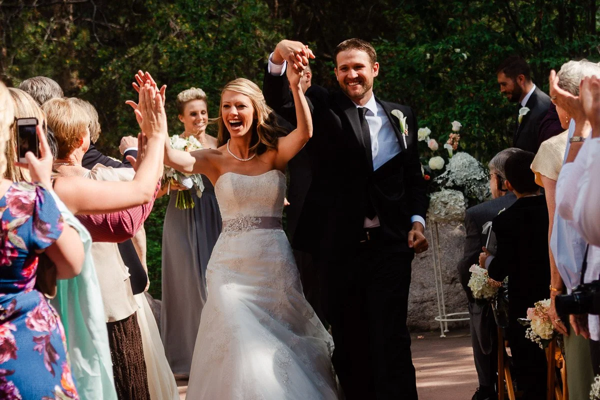 A joyful bride and groom walk hand-in-hand down the aisle, surrounded by cheering guests. The bride smiles widely, wearing a white dress; the groom is in a black suit.