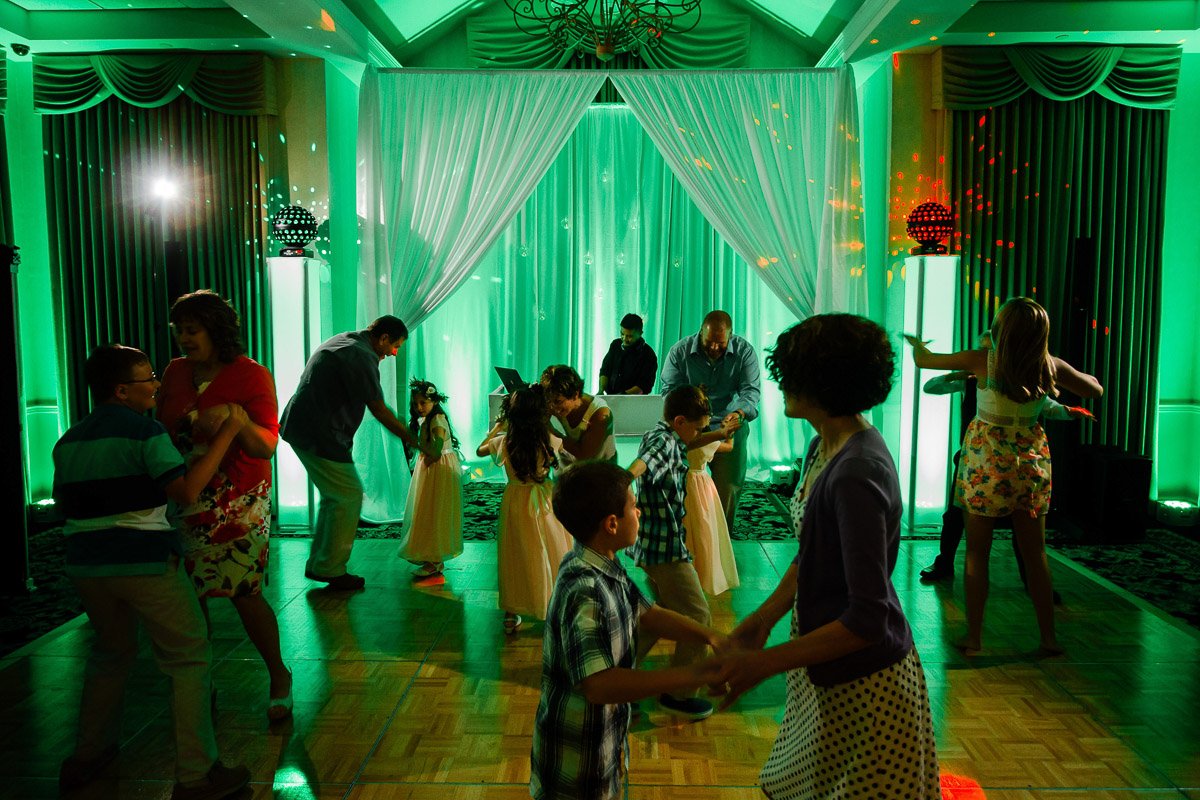 A lively dance floor at a family event, lit with green lights and drapes. Adults and children joyfully dance, creating a festive and energetic atmosphere.