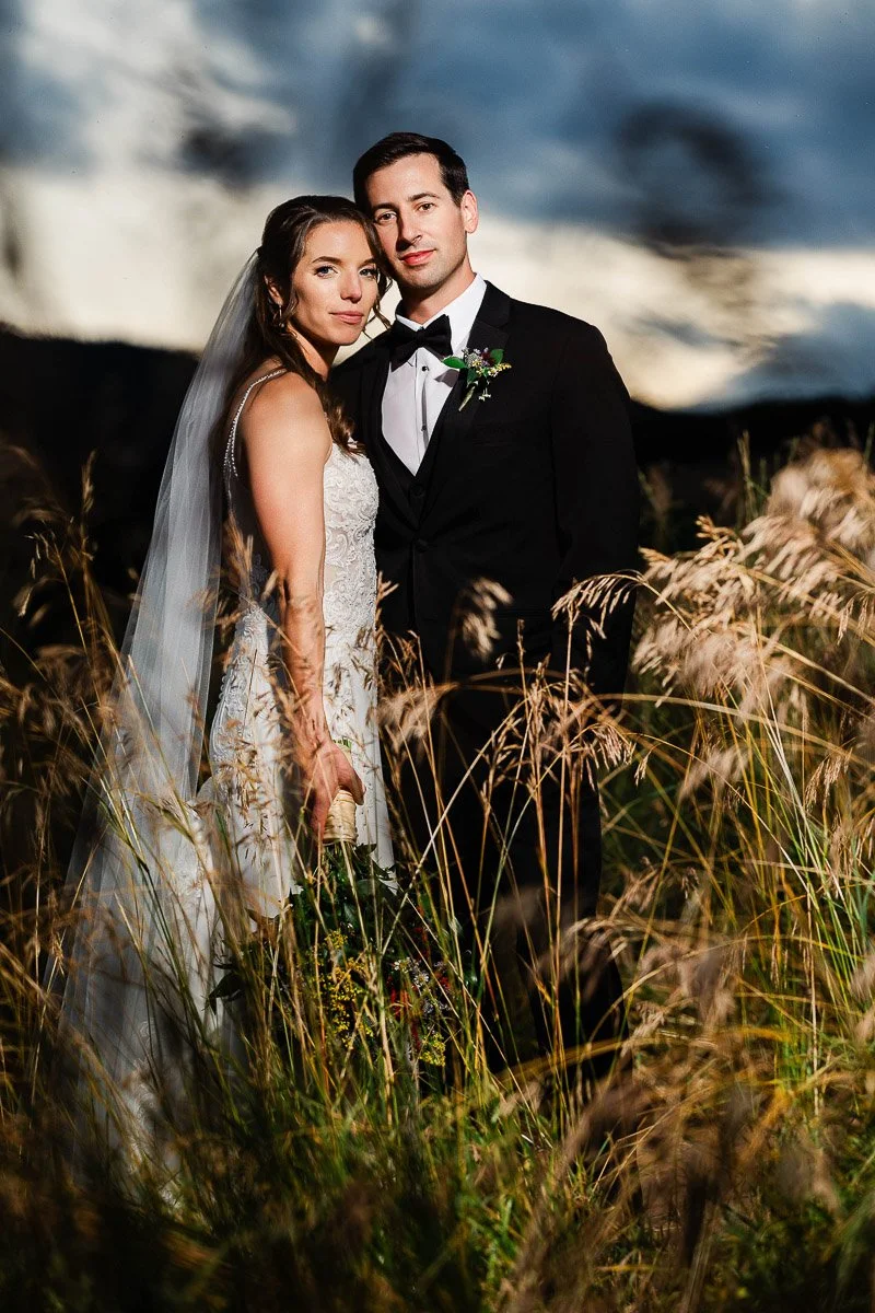 A bride in a lace dress and veil, and a groom in a black tuxedo, stand closely in a field of tall grass under a dramatic cloudy sky, conveying romance during a Spruce Mountain Ranch wedding in Larkspur, Colorado