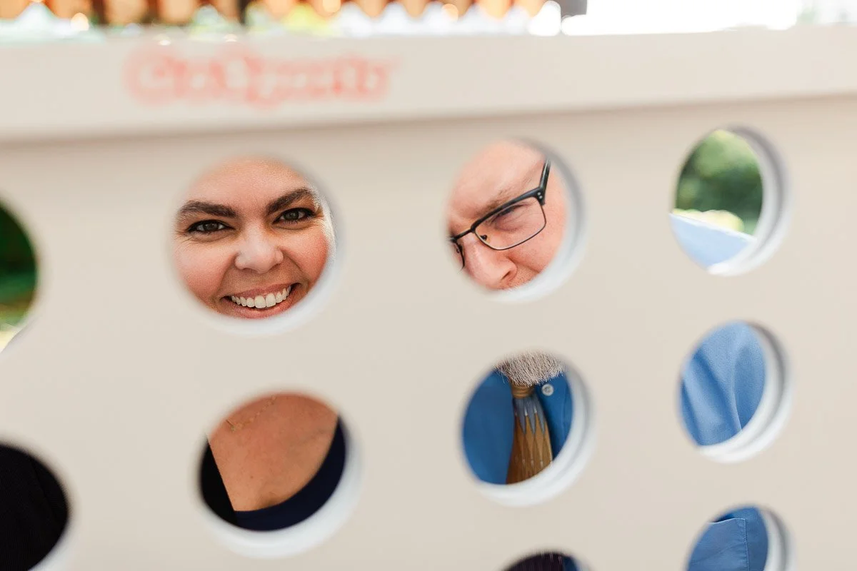 Two people laughing and seen through the holes of a giant Connect 4 game board, with a playful and joyful atmosphere in a sunny outdoor setting.
