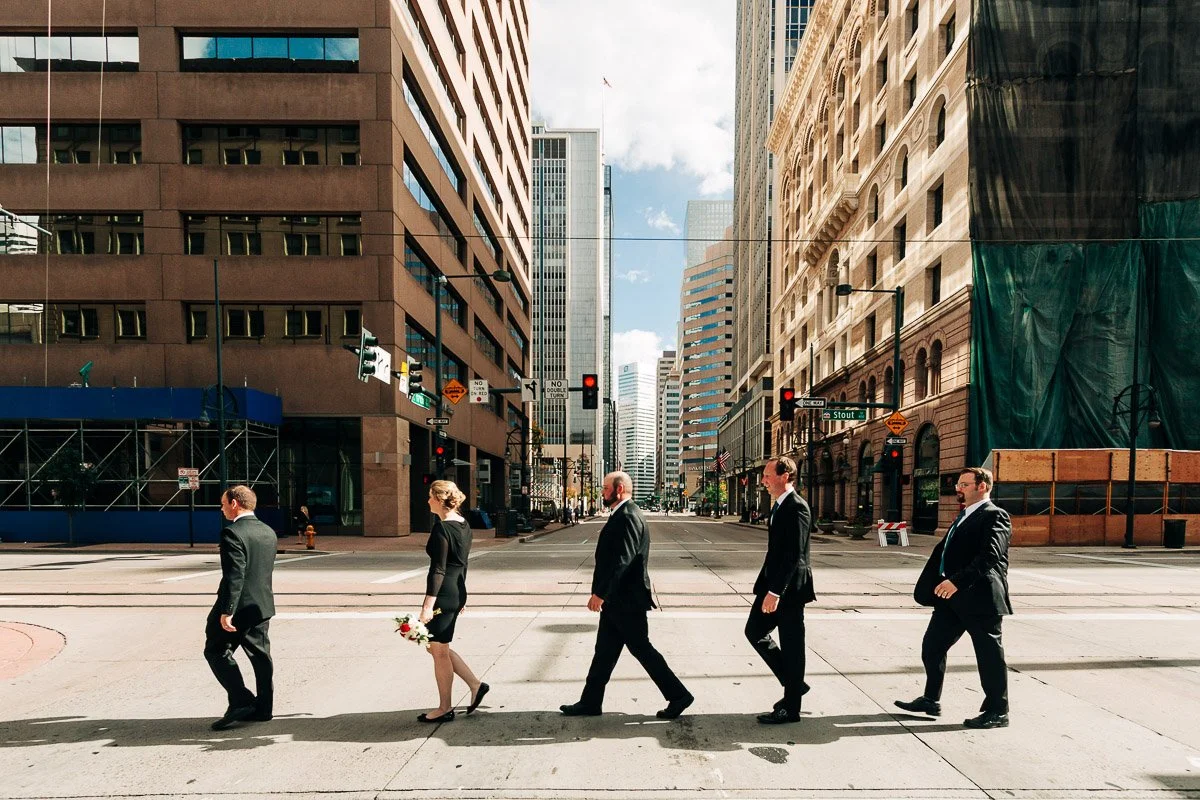Five people in formal attire walk across a city street, resembling a famous album cover. The atmosphere is lively, with tall buildings around them.