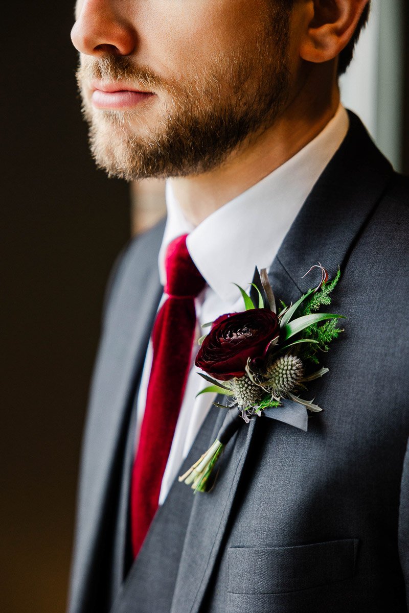 Man in a gray suit with a red tie, adorned with a dark red boutonniere against a neutral background, exuding elegance and sophistication.