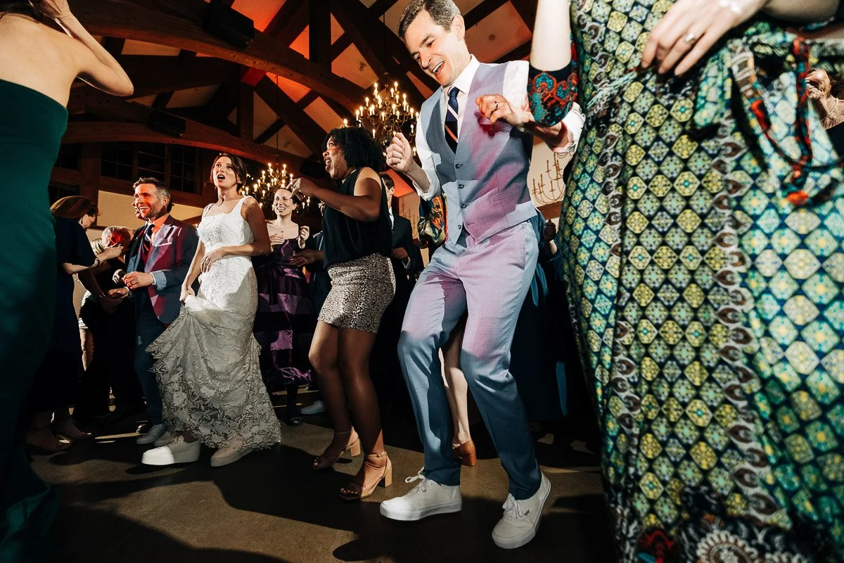 A joyful group dances energetically at a wedding reception. The bride in a white gown and guests in colorful attire celebrate under warm, elegant lighting.