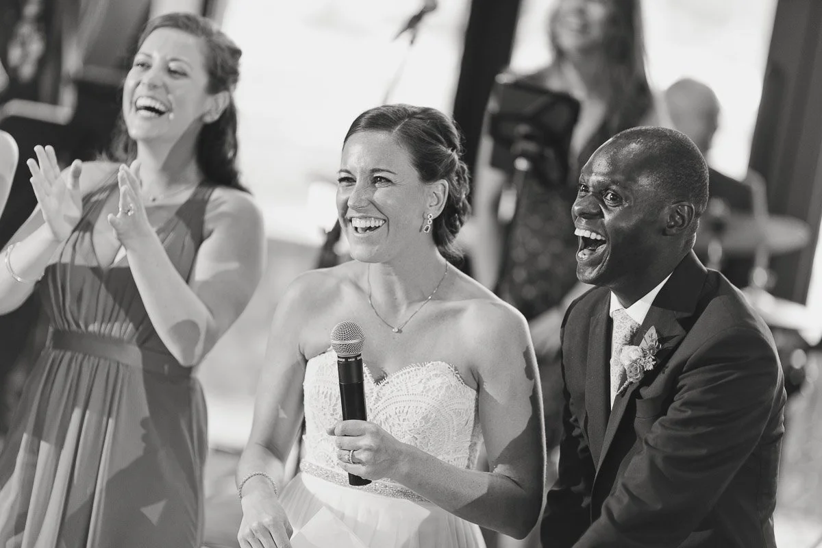 A bride holding a microphone, a man in a suit, and a woman in a dress share a joyful moment, laughing and clapping at a celebration. Black and white.