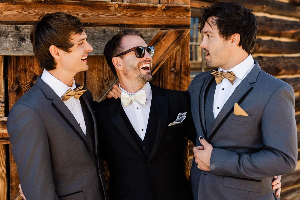 Three men in formal suits, two in gray with gold bowties and one in black with a white bowtie, share a joyful moment outside a rustic wooden building.