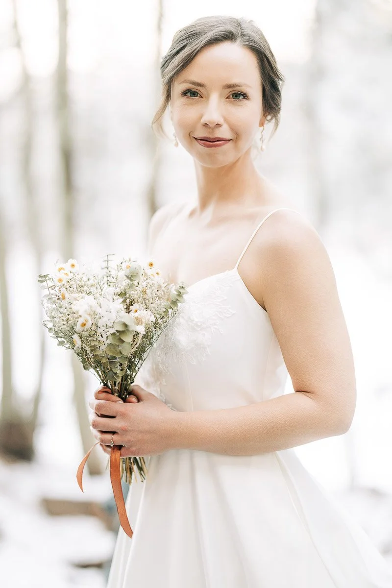 A bride in a white dress smiles softly, holding a bouquet of daisies and greenery. The background shows a snowy forest, creating a serene, elegant atmosphere.