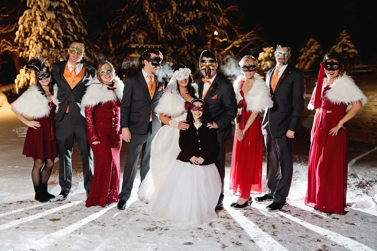 A group of people in formal attire and masquerade masks stand on snow at night, featuring two newlyweds in the center, creating a festive and elegant scene.