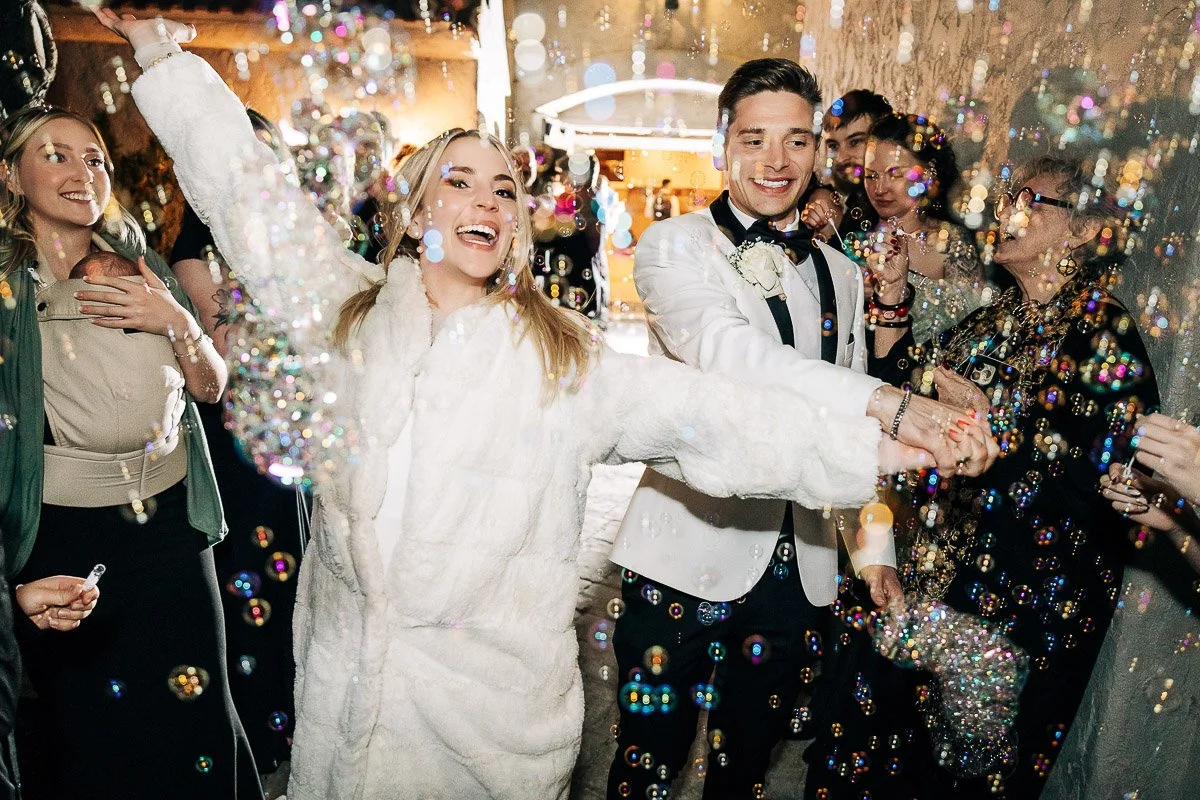 A joyful couple, dressed in formal wedding attire, walks through a shower of bubbles, surrounded by smiling guests. The scene is festive and lively.