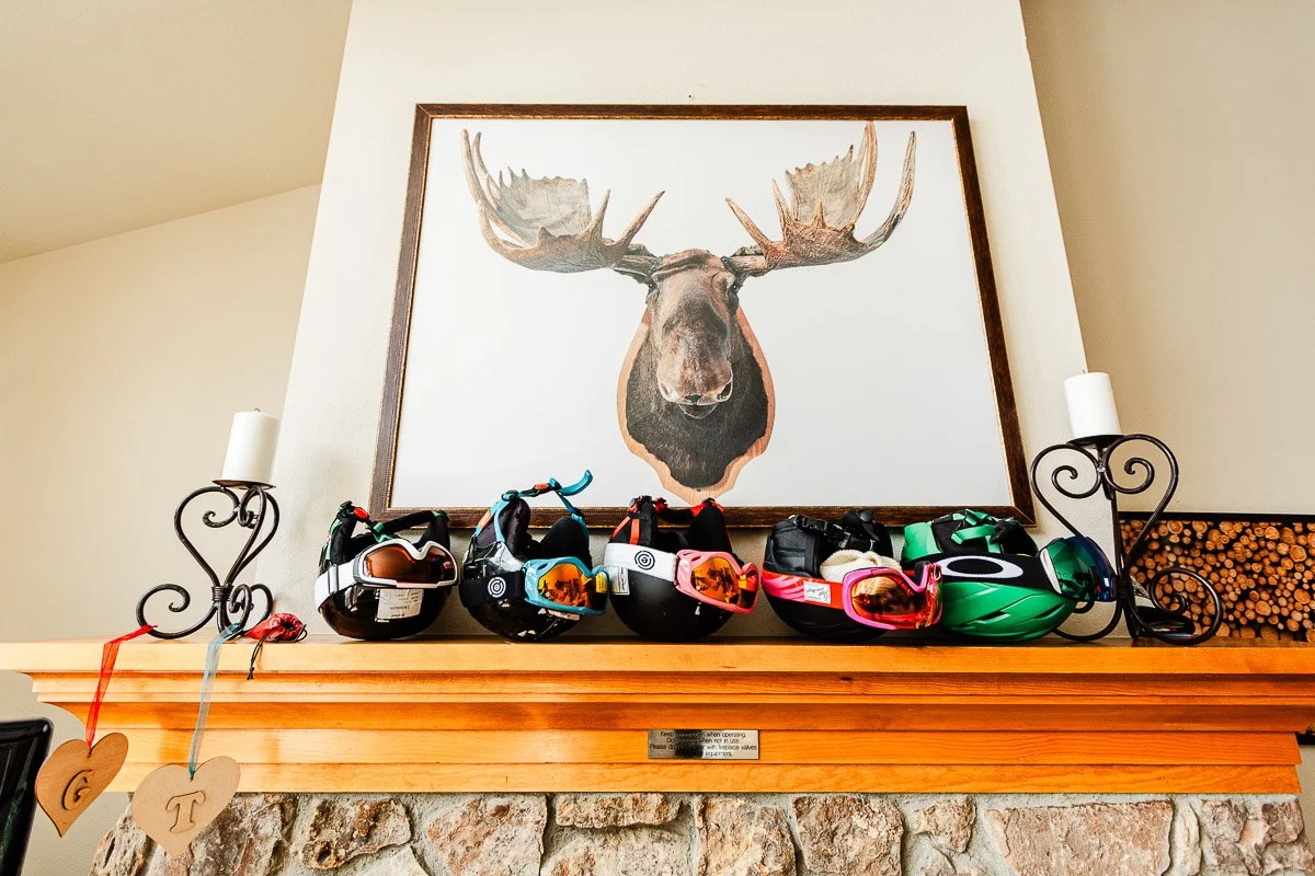 A framed moose portrait sits atop a rustic stone fireplace mantel, surrounded by ski helmets and goggles. Two candles and heart tags add a cozy touch.