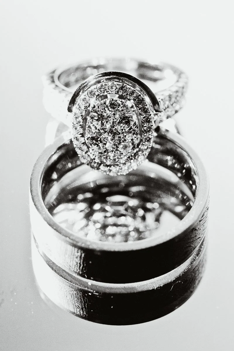 Close-up of two reflective rings, one with ornate diamond detailing and another smooth band, on a mirrored surface. Elegance and luxury radiate.