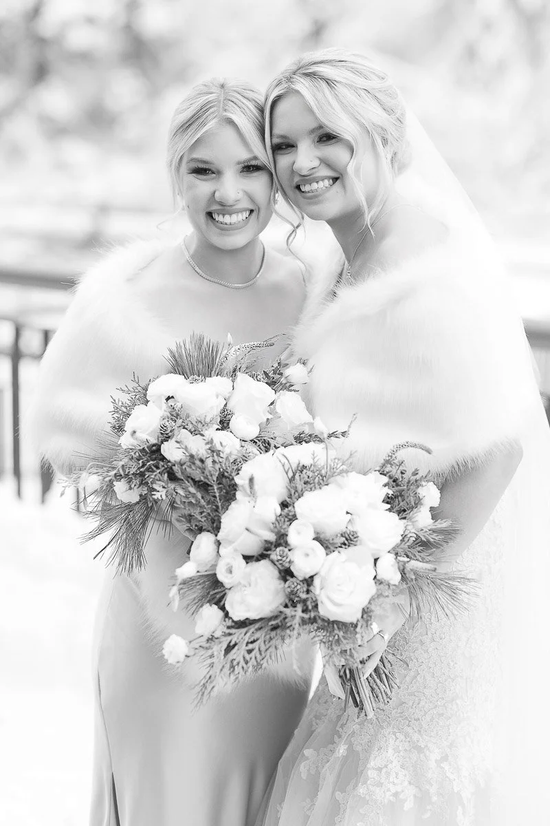 Two smiling women in wedding attire with fur capes hold snowy floral bouquets. The winter setting adds a joyful and elegant tone to the scene.