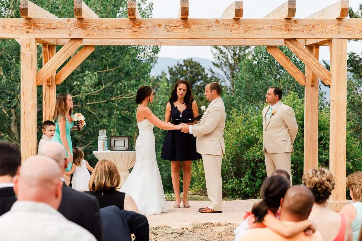 An outdoor wedding ceremony under a wooden pergola, with a bride in white and groom in beige holding hands. Guests are seated, and trees fill the background.