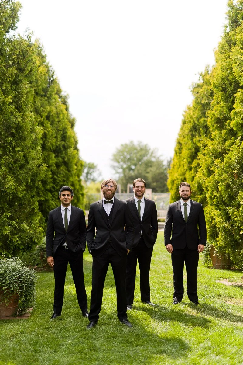 Four men in black suits stand on a neatly trimmed lawn lined with tall hedges. They appear confident and relaxed, suggesting a formal event at the York Street Denver Botanic Gardens