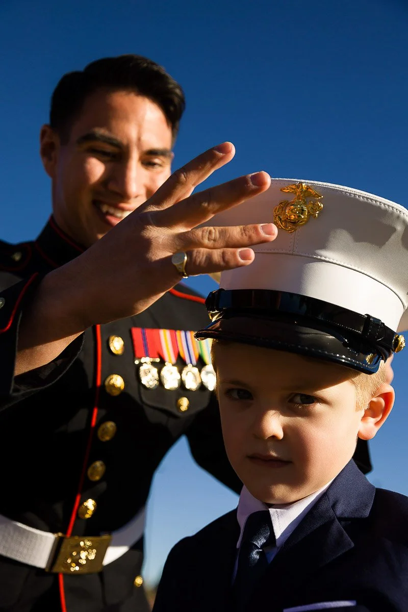 A smiling man in a Marine dress uniform adjusts a white military hat on a solemn young boy in a suit, set against a clear blue sky.