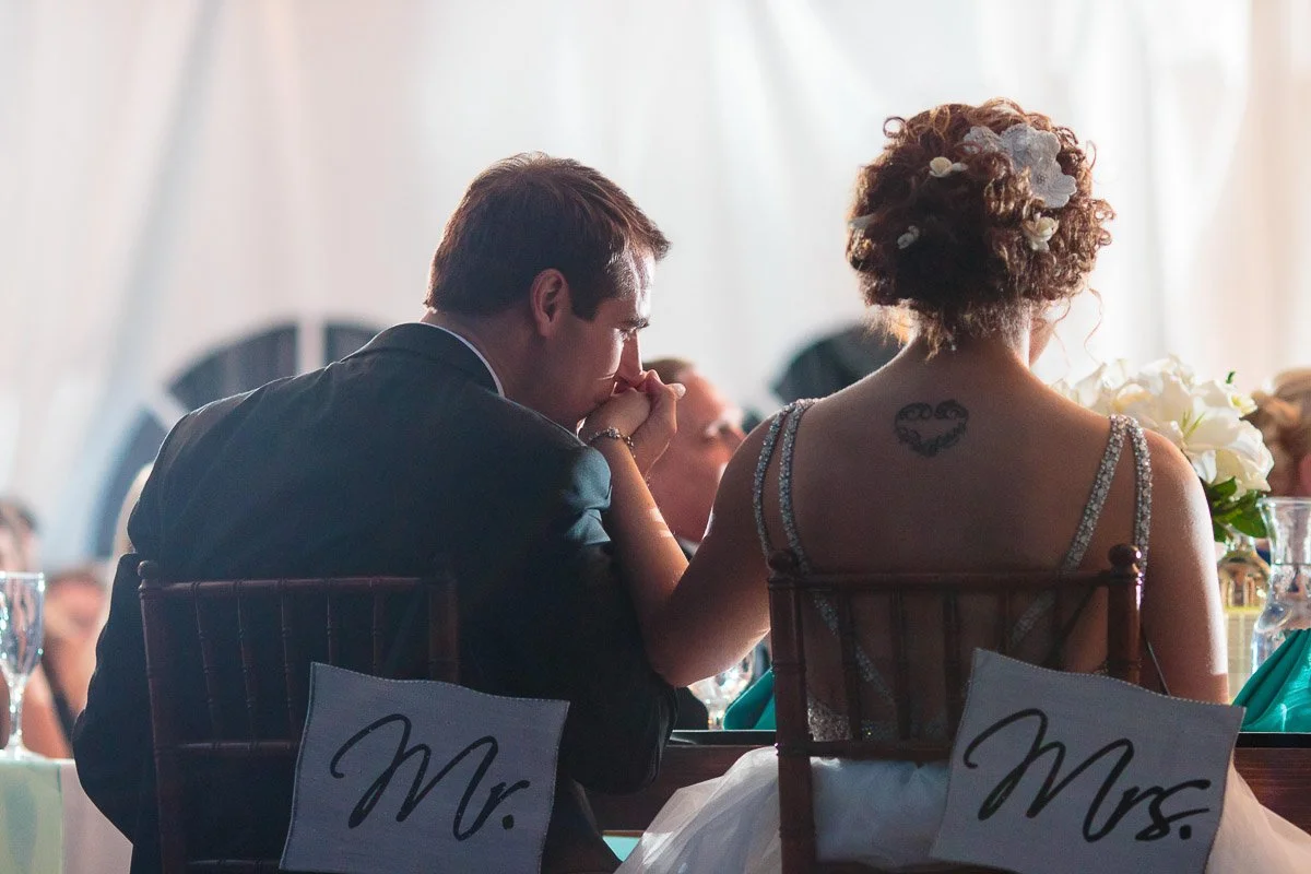 Wedding couple sits intimately at a table with "Mr." and "Mrs." signs on chairs. Bride's tattoo visible; soft lighting creates a romantic mood.