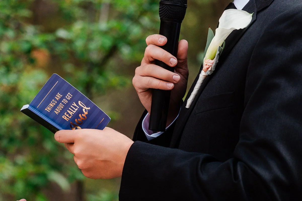 A person in a suit holding a microphone and a small notebook with the text "Things are about to get really good." A white lily boutonniere adorns their lapel.