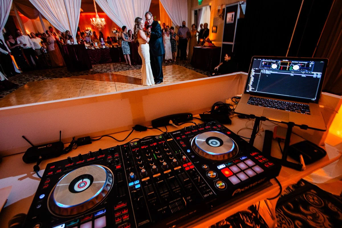 DJ equipment in the foreground at a wedding reception. A couple dances intimately on the wooden floor, surrounded by warmly lit guests in elegant attire.