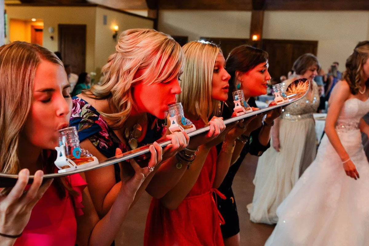 Four women at a party do shots from a ski shot board, laughing and enjoying. They are dressed in bright colors, conveying a fun and lively mood.