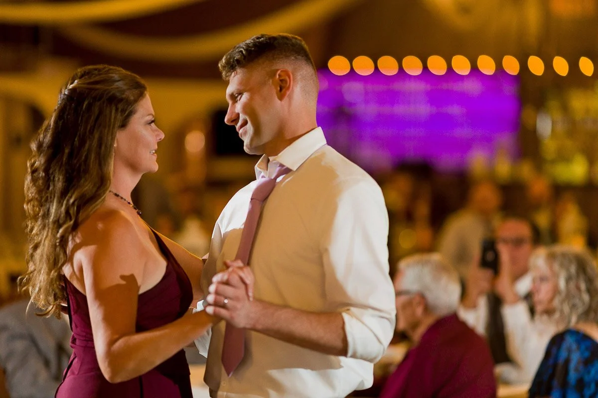 A couple dances closely at a warmly lit event. The woman in a burgundy dress smiles at the man in a white shirt and pink tie. Guests are blurred in the background.