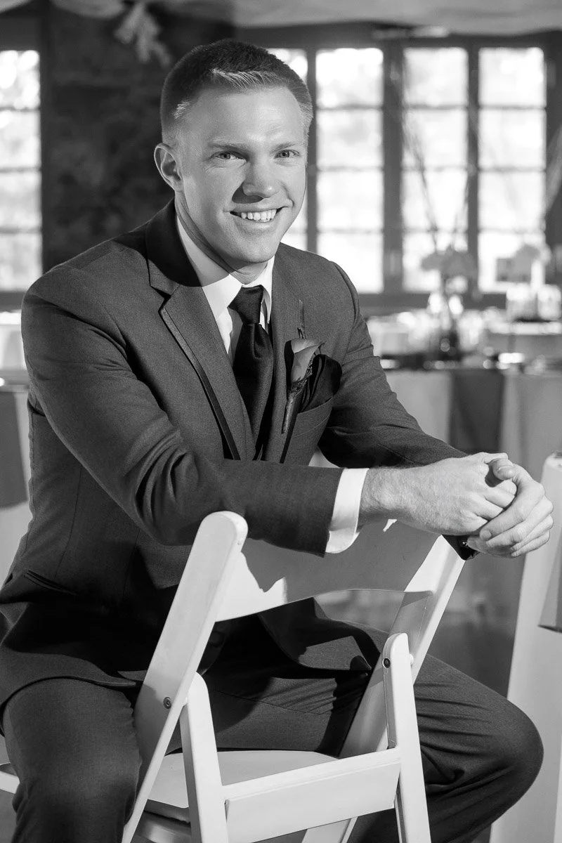 Smiling man in a suit sits on a chair in a sunlit room with large windows and round tables, conveying a warm and relaxed atmosphere.