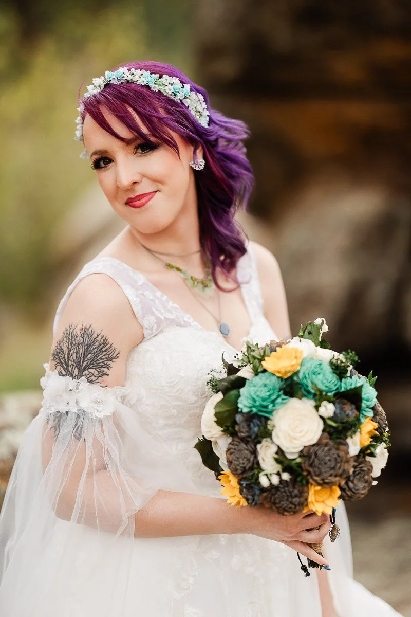 Bride with purple hair smiles warmly, holding a colorful bouquet with teal, yellow, and white flowers. She wears a lacy white dress and has a tree tattoo.