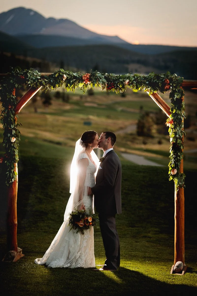 A bride and groom share a kiss under a flower-adorned wooden arch at sunset, with mountains and a grassy landscape in the background. Romantic and serene captured by Keystone wedding photographer tomKphoto
