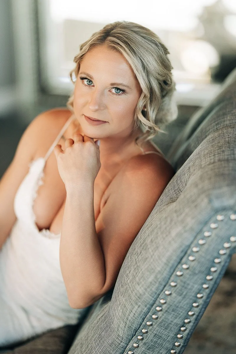 A woman with light hair sits on a gray upholstered chair, wearing a white dress. She looks thoughtfully at the camera, exuding a calm and serene mood.