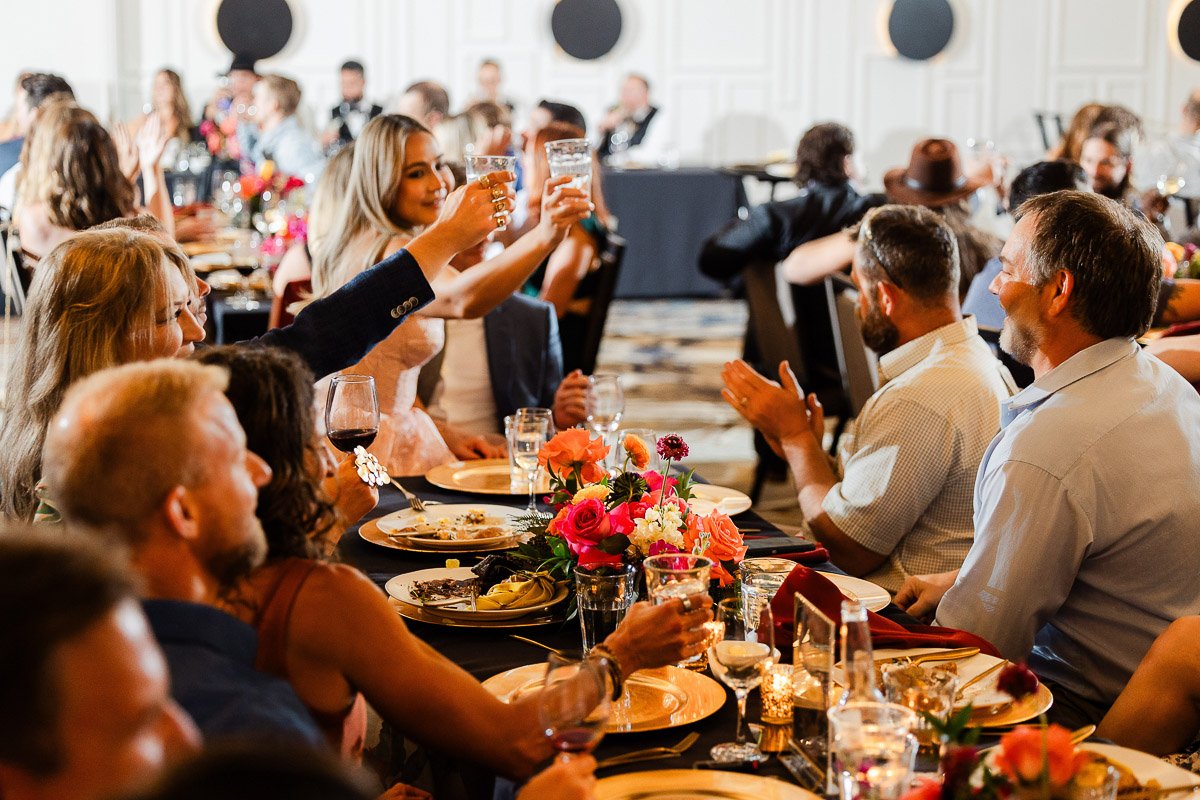 People gathered around a banquet table, raising glasses in celebration. Warm lighting, colorful floral centerpieces, and joyful ambiance.