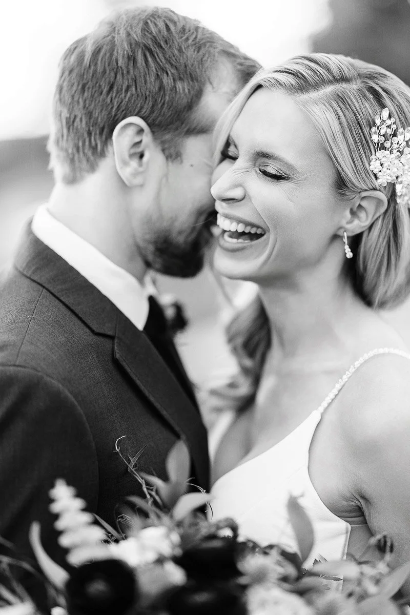 A black-and-white photo of a joyful couple on their wedding day. The woman laughs heartily, holding a bouquet, while close to her partner. Romantic and happy atmosphere.