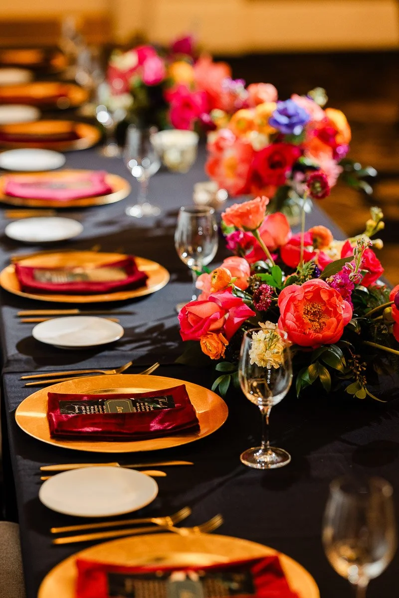 Elegant dining table with a black cloth, vibrant floral centerpiece of pink, orange, and purple flowers, gold place settings, and red napkins. Warm, festive tone.