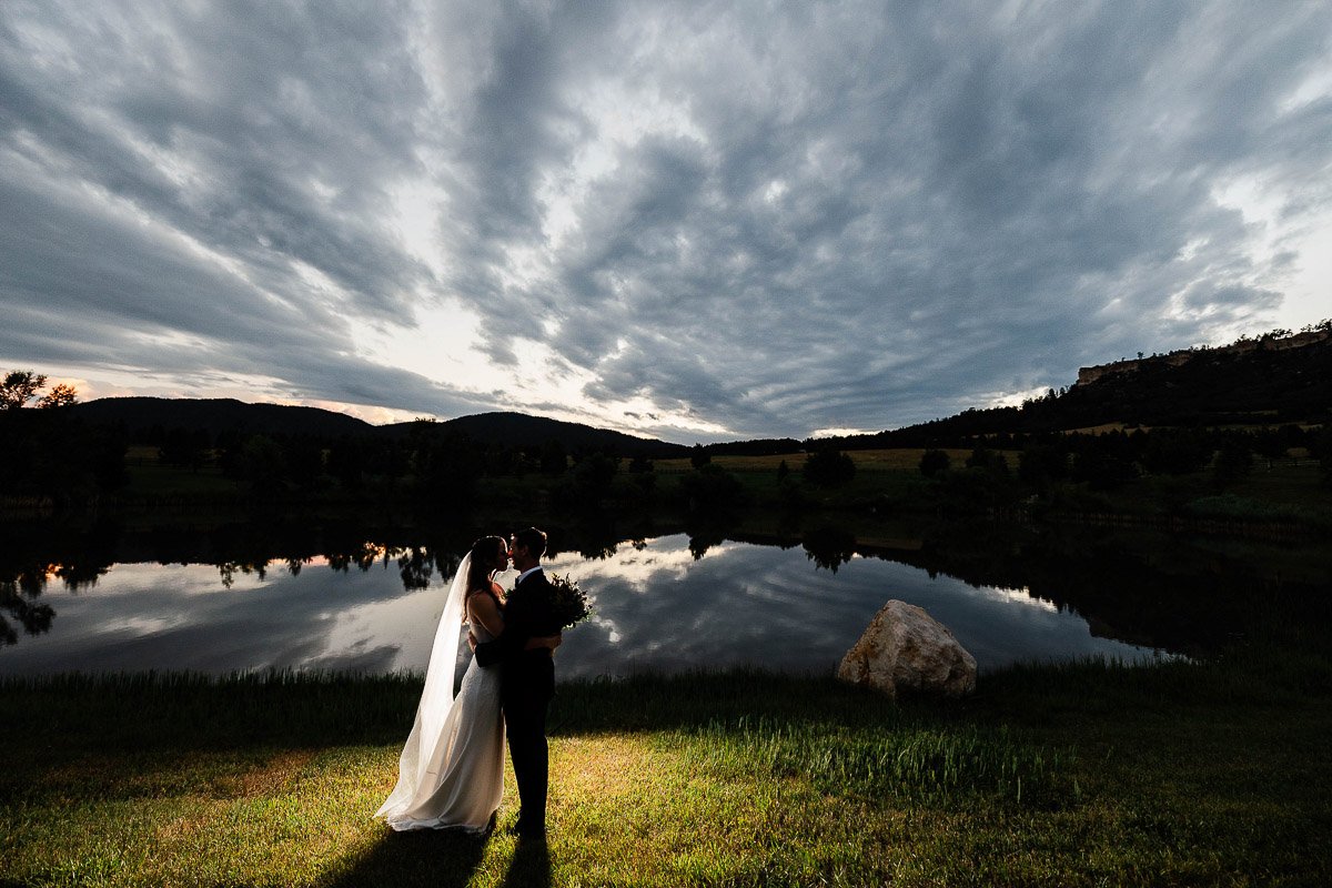 A bride and groom kiss in front of a serene lake and dramatic, cloud-filled sky at dusk. Their silhouettes are softly lit, creating a romantic and tranquil mood during a Spruce Mountain Ranch wedding in Larkspur, Colorado