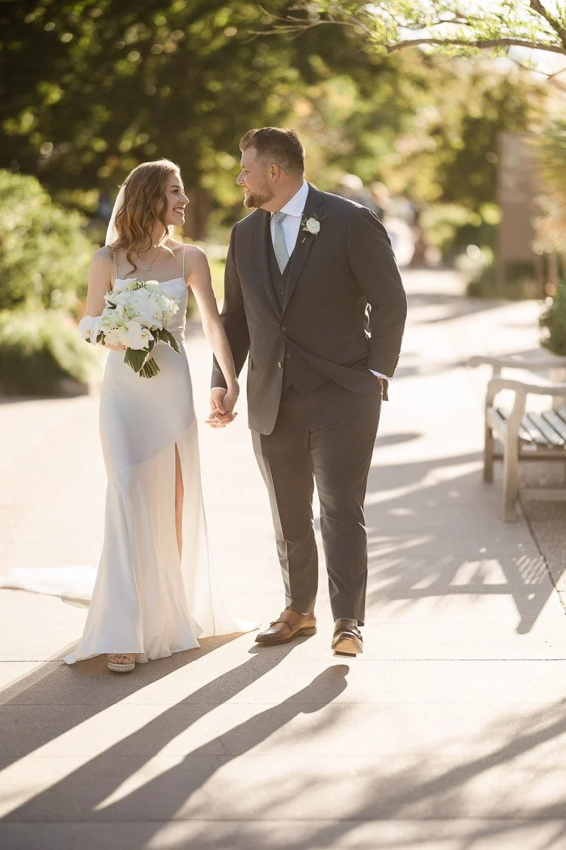 Bride and groom walking hand in hand on a sunny pathway, smiling warmly at each other. The bride holds a bouquet, and light filters through trees. Romantic atmosphere.