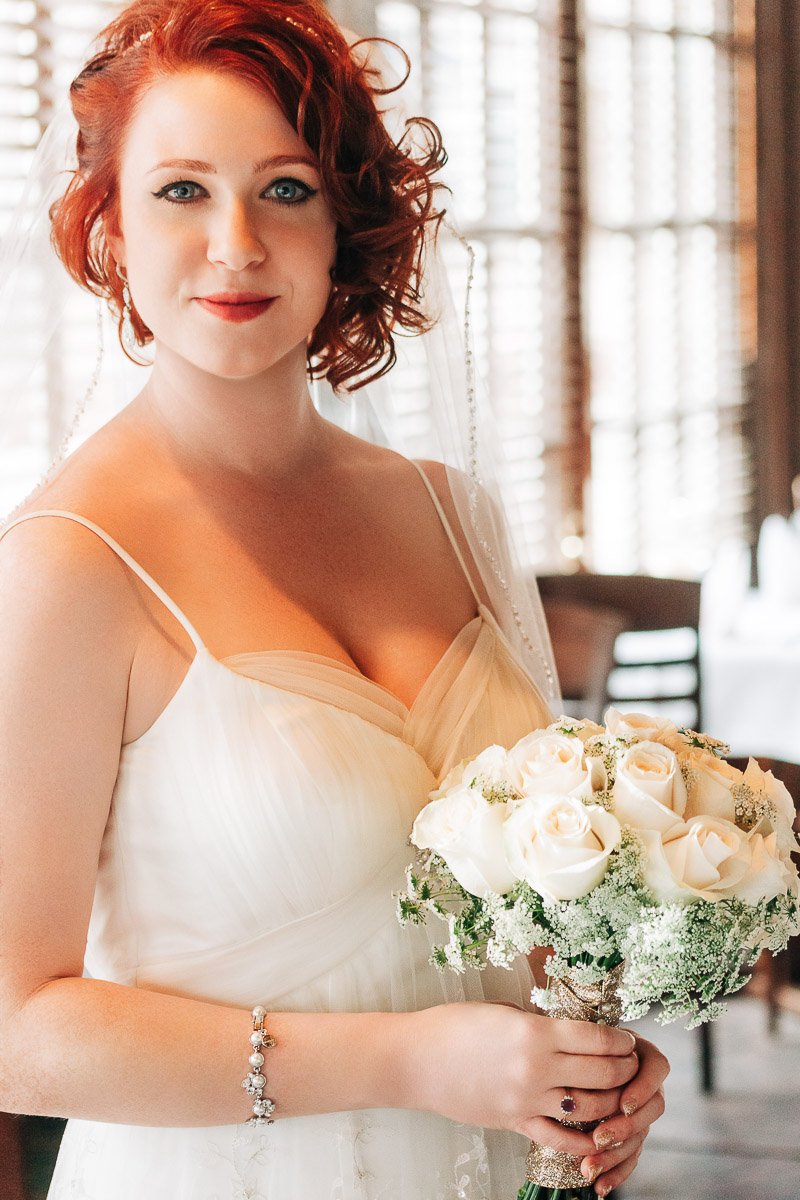 A bride with red hair smiles gently, holding a bouquet of white roses and greenery. She wears a white dress and a veil, creating a warm, joyful tone.