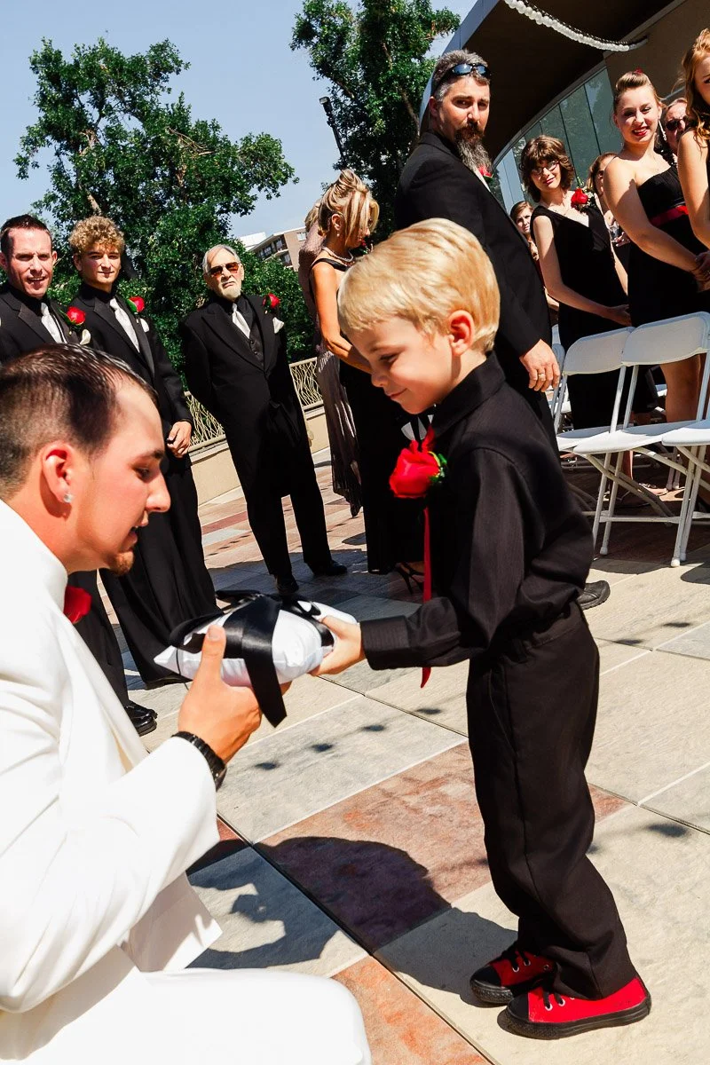 A young boy in black suit and red shoes hands a small pillow to a man in a white suit. They are outdoors, surrounded by smiling wedding guests.
