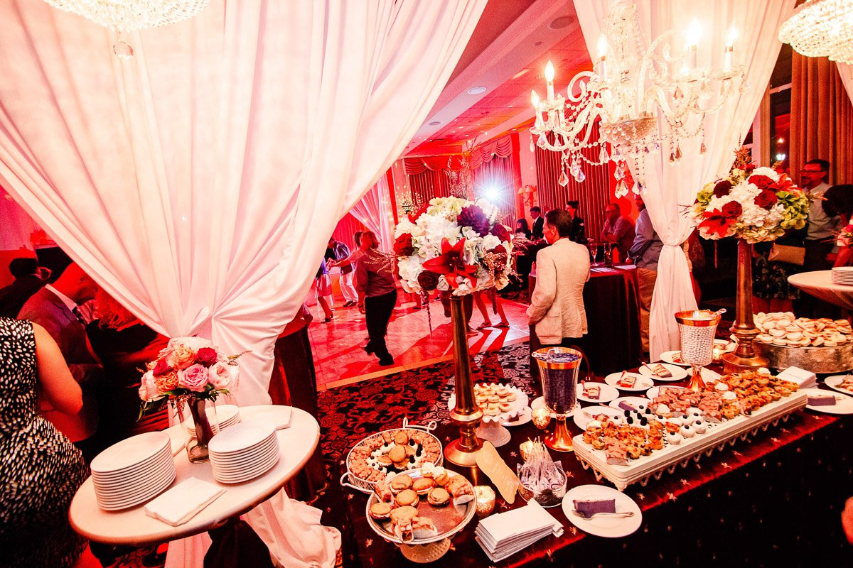 Elegant event with dim lighting, featuring a lavish buffet of desserts and flowers beneath chandeliers. Guests socialize in the background.